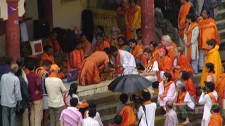 Outdoor photo of a ghat-side ritual on stone steps by a river. In the centre, a man in an orange robe kneels and bends forward over a small wooden stool, while a man in white with an orange shawl sits on a low seat beside him, and a woman in white reaches toward a brass object. Around them, devotees in orange and saffron garments sit on the steps and stand behind, with several women in orange shawls and white garments. A black umbrella shields part of the scene. Photographers in orange and white stand at the lower steps. A crowd of onlookers in casual clothing fills the left foreground, and a yellow banner with Devanagari script hangs on the wall behind.