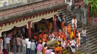 Outdoor photograph of a riverside temple complex with red-pillared arcades and stone steps. A large group of devotees in saffron and orange robes sits and stands on the steps and beneath the arcade, many wearing orange shawls. A man in a white kurta and orange shawl sits at the centre of the seated group, with a woman in white nearby. Around them, crowds of onlookers in ordinary clothes stand on the left and along the stairs, including men in shirts and trousers. A photographer with a camera stands on the steps at right. A carved stone figure sits on a pedestal in the upper right, and a sign with Devanagari text is on the wall. The scene suggests a gathering at a ghat.
