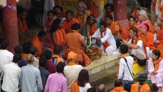 Outdoor photo at a riverside ghat with red stone pillars and a yellow painted wall. A crowd of people in orange and saffron clothing sits and stands around a central figure in an orange robe seated on a low step beside a small brass or bronze deity murti garlanded with white flowers. A man in a white kurta sits beside the murti, and a person in white and orange holds a microphone stand nearby. Several women in orange shawls and a woman in a pink headscarf sit in the group, with a bearded man in a white robe behind. Observers in white shirts and pink clothing stand at the lower edge of the frame, and a man with a camera bag is at right.