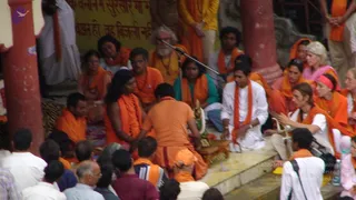 Outdoor photo on riverside ghat steps of a ceremony with a seated figure in orange clothing on a low wooden seat. Around the central area, many people wear orange shawls and saffron robes; a bearded man with a white beard and rudraksha-style necklaces sits near a microphone stand. A woman in orange and another in a white dress sit in the foreground, and a man in white with an orange scarf sits beside them. A red pillar and a yellow wall with Hindi script frame the scene. A crowd of onlookers in the lower foreground watches. The visible crowd includes more than twenty people.
