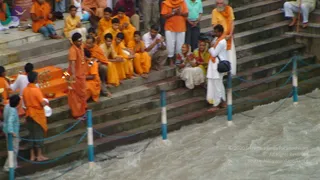 Outdoor river ghat steps beside fast-flowing brown water, with a blue rope railing and white-and-blue posts. A group of devotees in orange and saffron robes sits and stands on the stone steps, many with hands joined in prayer. In the left foreground a man in orange robes and a rudraksha mala holds a lit oil lamp and a brass ritual vessel near an orange-draped low platform. Behind him seated devotees wear saffron and yellow clothing, and a man in orange holds a small brass object. On the right a man in white kurta with an orange shawl stands at the water's edge, and two women in saris sit on the steps. A pale-clothed cameraman stands near the right, and a man in white on the upper right steps is partly visible. The scene suggests a riverside puja or aarti.