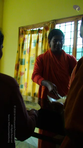 Indoor photo in a room with yellow walls and yellow floral curtains over a barred window. A young man in an orange-red robe wrapped over his shoulder stands at a table, reaching toward a plate with a spatula, apparently serving food. Two other people are partly visible at the edges: one in a dark maroon top on the left and one in an orange robe on the right. Their faces are not shown.