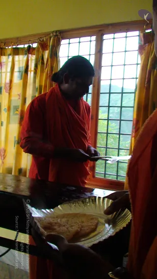 A man in a saffron-orange robe and matching wrap stands at a dark wooden counter beside a window with grey metal grilles and yellow floral curtains, with green hills outside. He holds a spatula and serves a crisp dosa onto a large round silver-toned plate held by a second person in orange at the right edge of the frame. The man has long dark hair tied back, and the scene appears to be a kitchen-style serving area in an ashram setting.