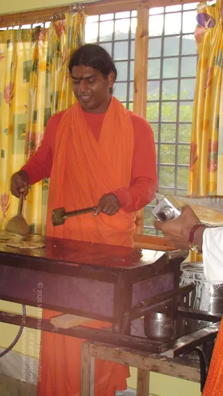 A photograph of a man in an orange saffron wrap over a red-orange top standing at a wooden counter, spreading a dosa batter on a flat griddle with a metal ladle-like spreader in one hand and a flat spatula in the other. Yellow curtains with floral patterns hang at a window with grid bars behind him, showing green trees outside. Metal vessels sit on the counter, and another person's arm in a white sleeve holds a camera at the right edge. The man has long dark hair swept back and a tilak on his forehead, and he smiles as he cooks.