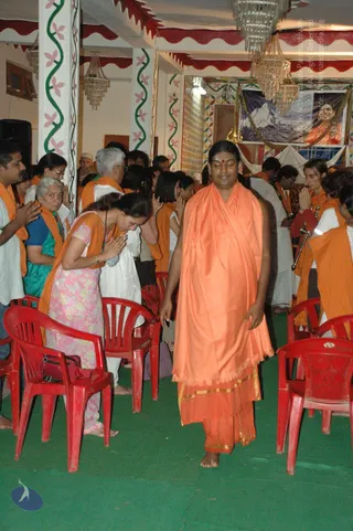 A photograph inside a decorated hall with green floor, red plastic chairs and chandeliers. In the centre a man walks barefoot in a flowing saffron-orange shawl over an orange dhoti with gold borders and a red tilak on his forehead. Devotees on the left and right stand with palms pressed together in anjali, some in orange shawls, a woman in a pink floral salwar and an older woman in teal sari among them. A banner on the wall shows a snowy Himalayan peak and a portrait of a smiling man in orange robes, with partial text reading Nithyananda and 2008 printed on it. Two people in the background hold ceremonial items.