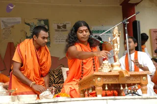 A photograph of a ceremonial puja at an outdoor platform. A man with long, swept-back dark hair and saffron-orange robes, wearing a rudraksha mala, sits at a carved wooden altar and gestures toward a brass deity murti and a stack of small offerings. Beside him, a younger man in orange robes sits attentively with a silver vessel and brass bowls in front of him. Behind them, a young man in white with an orange shawl sits with a microphone stand nearby. Hanging bells decorate the altar's edge, and marigold flowers and a small lamp rest on the platform. The wall behind shows faded posters and a painted map.