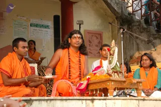 A photograph of a ritual on an outdoor stone platform beside a weathered yellow wall with pasted notices and a carved red sandstone relief. A central figure in saffron-orange robes with long swept-back hair, rudraksha malas and a tilak sits behind a carved wooden table draped with orange marigold garlands, holding a small plate. A deity murti with a jeweled crown and pearl garland stands on the table beside a bell-hung stand. A saffron-clad attendant in an orange shawl sits to the left holding a plate, another man in white sits behind the murti, and a woman in a teal top and orange shawl sits at the right edge, with a microphone on a stand nearby. Several spectators watch from a metal railing above.