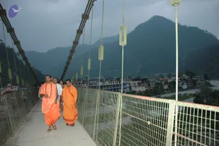 A photograph of a suspension footbridge with a grey mesh railing and steel cables, crossing a river at dusk. Two men in saffron-orange robes walk toward the viewer on the bridge deck. The man on the left wears a rudraksha mala and has long dark hair and a tilak on his forehead; the man beside him wears a saffron wrap over his shoulder. Behind them a few people in white and light clothing follow. Hills rise behind a town of multi-storey concrete buildings along the riverbank, under an overcast sky.