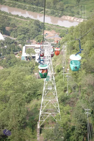 A photograph of a ropeway cable car system descending a steep, densely forested hillside. Several enclosed cabins in green, red, orange and turquoise hang from a steel cable strung between white lattice towers. A muddy brown river runs along the top of the frame beside a green embankment, and a white building with a flat roof sits at the upper left among trees. No people are visible.