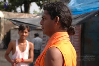 A photograph of a man in profile wearing a sleeveless saffron-orange garment wrapped over his shoulder, with dark hair tied in a bun at the back of his head and a light vertical mark on his forehead. Behind him, a young man in a white vest with a red wristband smiles, and a blue tarpaulin and dark shade cloth stretch over a brick structure in an outdoor setting.
