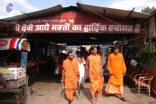 A street scene at a market arcade beneath a red banner with white Hindi lettering and a saffron-coloured Sanskrit invocation above. Three men in saffron and orange robes walk left to right; the central figure, with long swept-back hair and a dark shawl over his shoulder, carries nothing in hand, while the man at right wears an orange robe and a beaded necklace. Behind them a person in white clothing carries a speaker box, and a child in white walks nearby. Stalls with ice cream signage, plastic chairs and produce crates line the arcade.