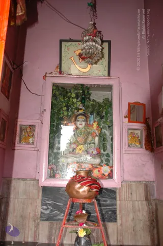 A shrine niche in a pink-washed wall holds a white marble deity murti seated in padmasana, draped with fabric and flower garlands in orange, yellow and pink, set against a backdrop of green leaves and dark berries. A framed Om symbol hangs above with a shell-and-cord ornament. Small figures in yellow garments flank the main shrine. In front stands a copper kalasha wrapped in a red and white striped cloth on a red metal stand, with flowers and a black vessel at the base. No people are visible.
