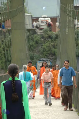 A photograph of people walking across a narrow suspended footbridge with wire-mesh side railings, toward a parked white van and roadside buildings with green hillside behind. In the foreground a woman with a long side-braid wears a navy top and bright green shawl, seen from behind. Beside her, a woman with grey hair wears a turquoise top and carries a white shopping bag. Further ahead, several men in saffron-orange robes walk together; one has long dark hair and another has a grey beard, and a boy in a pink shirt and jeans walks in front of them. A man in a blue polo shirt and brown trousers walks at the right edge of the bridge. The people walk on a gravel path, and the scene reads as an outdoor journey in hill country.