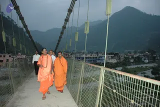 A photograph of a suspension footbridge with metal mesh railings and cables, crossing a river valley at dusk with wooded hills and a small town behind. In the foreground, a bald man with a shaved head and a rudraksha mala wears a saffron-orange robe and walks in sandals. Beside him, a second man in an orange robe with a red border walks along the bridge. Behind them, two men in white shirts and dark trousers follow across the walkway.