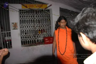 A photograph inside a narrow cave-like passage beside a barred metal grille window with a shrine behind it. A man in long flowing saffron-orange robes and a long rudraksha mala stands at center, with long dark hair swept back and a red bindi on his forehead. Two partial figures appear at the edges, one in the foreground right in a white shirt and one at the left edge. A red box marked donate box in Hindi and English sits below the grille, a painted image of Hanuman is on the wall at left, and a yellow banner reading Om Namah Shivaya is hung above with colourful garlands strung along the wall.