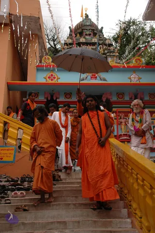 A photograph of saffron-robed devotees descending a concrete staircase in front of a colourful South Indian style temple gopuram with a painted blue facade. In the foreground a bearded man in orange kurta and dhoti walks barefoot, and beside him a figure with long dark hair wears an orange robe and a long rudraksha mala, one hand holding a black umbrella overhead. Behind them other devotees in white dhotis and orange shawls follow, and an older bearded man in white with a rainbow garland stands at right near a deity murti shrine. Shoes are lined up on the steps at left, and a yellow sign reads 'Remove Your Footwear Here'. Streamers and tinsel hang from the rooftops above.