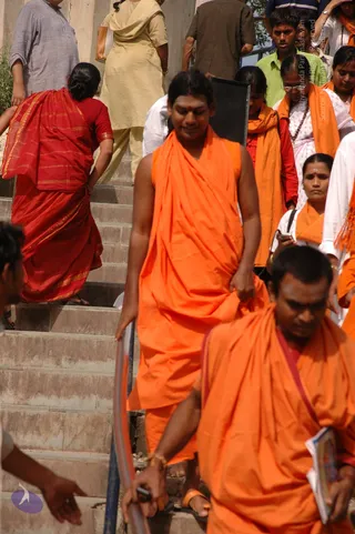 A photograph of a crowd descending concrete steps outdoors in bright sunlight. In the centre, a young man in a loose orange sleeveless robe with a red mark on his forehead walks down the steps, one hand on a metal railing. Beside and below him, a man in orange robes holds a small booklet. Around them, women in orange shawls and saris, a woman in red, and men in plain shirts stand or climb the steps. Several people are visible behind, including one with a mobile phone.