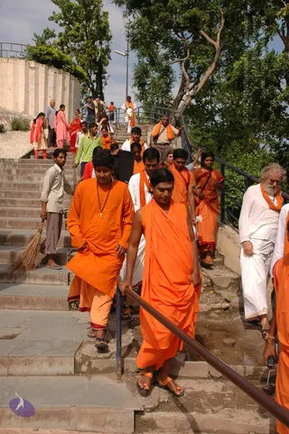 A photograph of a group descending a stone staircase outdoors beneath trees, with a metal handrail on the right. Several people in saffron-orange robes and shawls walk down in a line. In the foreground a young man in an orange robe and orange sandals holds the rail, and behind him a man in an orange kurta wears a rudraksha mala and pendant. On the right an older bearded man in a white kurta and white dhoti wears an orange shawl. Other people in orange and white clothing follow, and a man in a green shirt and a younger man in grey trousers holding a broom stand on the steps above. A few people in pink and red salwar stand on the upper landing near a concrete wall.