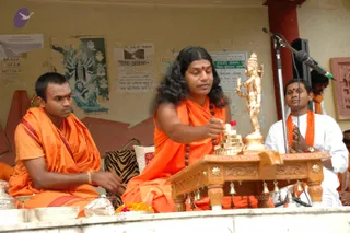 A photograph of an outdoor ritual on a raised platform against a weathered yellow wall with pasted posters. A central man in orange robes and a long rudraksha mala with long, wavy hair holds a flower over a small stepped brass pyramid beside a golden standing deity murti with raised arm. Orange-robed man sits to the left with a wrist band, and a man in white with an orange shawl sits with hands folded to the right. A carved wooden puja table with hanging bells and a silver plate of offerings sits in front, with a microphone on a stand behind. Possibly a puja or abhishekam.