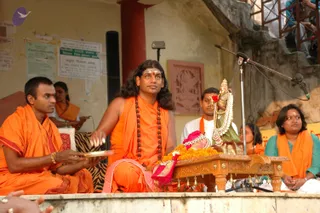 Outdoor ritual on a stone platform beside a yellow wall with posted notices. A man with long swept-back hair, a tilak on his forehead, wearing an orange shawl and a rudraksha mala, sits centrally before a carved wooden stand holding a decorated deity murti garlanded with orange marigolds and a red rose, with gold jewellery. A man in orange at left holds a brass plate. Another man in white and orange sits behind, and a woman in a teal top and orange shawl sits at right. Bells hang from the wooden stand and a microphone stands at right. Visible long-haired figure with rudraksha and orange clothing: a person, seated as a ritual presenter.