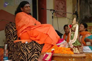 A photograph of a seated man in an orange saffron shawl and orange dhoti sitting in a tiger-striped armchair on a stage with a microphone on a stand behind him. He has long dark hair swept back, a tilak on his forehead, and his legs are crossed with one foot resting near a brass tray of marigold garlands and a pink cloth. Beside the tray stands a deity murti dressed in a green and gold sari with a gold crown and a layered pearl and gold necklace. Two women sit to the right, one in an orange shawl and one wearing glasses in an orange shawl, with a water bottle at the lower left.
