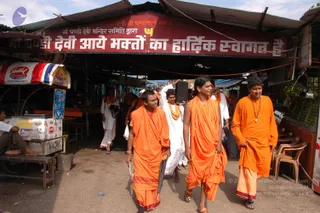 A photograph of four men in saffron-orange robes walking through a market area beneath a red banner with Hindi text. The man in the centre wears an orange robe and a shawl draped over his shoulder, while the figures beside him wear orange and saffron clothing; one holds a folded umbrella. Behind them, people in white clothing and orange shawls move through the crowd near an ice cream stall and fruit stall with green coconuts. A black speaker is visible on a pole.