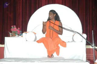 A man with long swept-back dark hair and a red bindi sits cross-legged on a large white cushioned throne-like seat before a deep red velvet curtain. He wears a saffron-orange sleeveless robe and a long rudraksha mala with a ceremonial necklace, smiling and gesturing with both hands while speaking into a gooseneck microphone. A white bolster cushion sits at his side, a small table holds a vase of pink flowers, a water bottle and a brass item, and white sheeting covers the platform in front. The appearance matches the Swamiji (probable) cues of long hair, saffron clothing and rudraksha mala, as a speaker giving a discourse at the Nithyananda Convention, Bidadi.