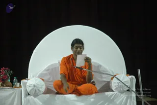 A seated man in orange saffron robes sits cross-legged on a white-draped raised seat in front of a large white circular backdrop and dark curtains. He holds a small white paper in one hand and gestures with the other, with a gooseneck microphone bent toward him. A vase of pink flowers and a water bottle sit on a white-covered table at left. The man wears a tilak on his forehead and has his hair swept back. Appearance-based label: a person, seated on the stage throne, wearing saffron robes and a sweeping hairstyle, giving a talk from written notes.