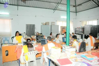 Photograph of a busy office-like hall with a light corrugated metal roof and a green painted support pillar in the centre. Several people sit at desks working on laptops and desktop computers, many wearing white clothes with orange or saffron shawls and sashes; one woman at left wears a white outfit with a yellow-orange shawl, and a woman in the foreground wears a yellow top with white garment. Stacks of books and printed booklets cover the tables in the foreground, and grey metal cabinets line the walls. The room has white walls and a few plastic chairs.
