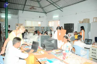 A photograph of a busy computer lab inside a plain white-walled building with a corrugated metal roof and ceiling fans. Devotees in white and saffron-orange clothing work at desktop monitors and laptops along tables with yellow cloths, books and a small bottle of water. A woman in a yellow top leans over a laptop on the left, while a woman in an orange shawl and rudraksha-style beads stands at the centre. Shelves of books and metal lockers line the back of the room, and a small carved figure sits on a table.