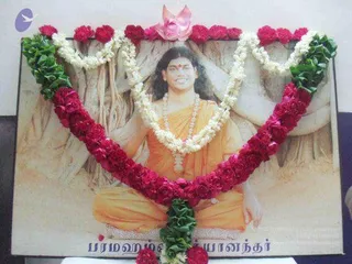 A framed printed portrait of a seated man in saffron-orange robes with a rudraksha-style necklace, bare-chested with a red tilak mark, set against a tree-root backdrop. The portrait is decorated with a horseshoe-shaped garland of red roses and white jasmine strands, with green leaves along the lower edge and a pink lotus-like flower at the top. Tamil script in the lower right corner of the print reads Paramahamsa Nithyananda, attributed to the caption on the printed portrait. Since this is a printed portrait rather than a live person, the subject is shown as a depiction.