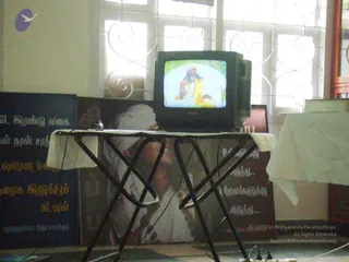 A photograph of an indoor room where a small CRT television sits on a folding table draped with a white cloth. The television screen shows a colourful animated or illustrated scene of a bearded figure in white with a child. Behind the table is a large printed banner showing a figure in white robes with a white head covering, next to Tamil text on dark blue and brown panels. Window grilles and a white wall frame the setting. No physical people are visible.