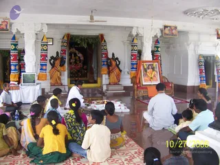 Indoor temple hall with white pillars and arches, decorated with striped colourful cloth-wrapped pillars. In the centre, a priest in white clothing performs a puja at a floor arrangement of white packets, brass items and a lit oil lamp. Seated devotees on red and patterned mats watch, including women in yellow, green, and floral saris and children in checked shirts. Behind, a framed portrait on a red-and-orange draped chair is garlanded with marigolds, showing a figure in orange robes; this is a printed portrait, not a person present. Two dark stone-coloured statues in orange dhoti-like garments flank a shrine, and a television sits on a table at left.