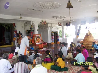 A photograph inside a white pillared hall with a polished floor, where seated devotees sit on mats while a standing man in a white shirt with an orange-trimmed collar tends a small fire near the front. Behind them stands a framed portrait of a long-haired figure in orange garlanded with flowers, placed on a red cushioned chair draped in orange cloth. A decorated deity murti stands at left, and a golden stepped structure stands at right. Colourful striped pillar wraps, a hanging bell and ceiling fans complete the room. Seated people include women in saris and children in yellow, pink and green clothing. The room holds about two dozen visible people.