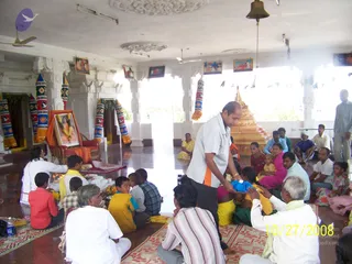 A photograph inside a white hall with pillars and a ceiling medallion, where many people sit on red and patterned floor mats. In the foreground a man in a white shirt with orange trim stands and hands a folded blue cloth to a seated child in yellow. To the left a framed portrait of a deity with garland sits on a red chair draped in orange cloth. A golden stepped shrine stands at the centre-right, with a hanging bell above it. Women in saffron, pink and yellow saris, children and older men in white sit among the devotees, and decorative striped cones hang from the pillars. A date stamp in the corner reads 10/27/2008.
