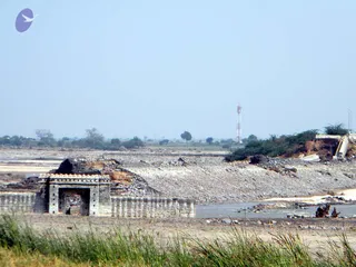 A wide photograph of a flood-damaged rural landscape under a pale hazy sky. In the left foreground a small stone gateway with arched opening and crenellated parapet stands amid collapsed masonry and debris. Behind it a broad expanse of grey gravel and silt covers the ground, with a river channel of shallow water running across the middle. On the right a broken concrete structure and an eroded earth bank lie beside a line of green shrubs. In the distance a red-and-white lattice telecom tower and scattered trees sit on flat farmland. Tall green grass fills the bottom edge of the frame. No people are visible.