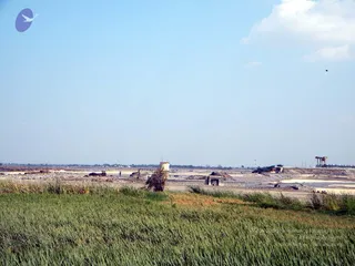 A wide daytime photograph of a flood-damaged landscape under a pale blue sky with a few clouds. In the foreground, tall green grass and reeds fill the lower frame. Behind them lies a broad expanse of cracked, sandy and muddy ground with shallow pools of water. A breached or broken dam structure sits across the middle distance, with a damaged concrete pillar or tower standing in the centre and a sluice-like gate section with metal railings to its right. A tall gantry or lifting structure stands at the far right edge. No people are visible.