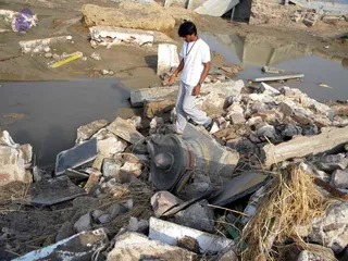 Photograph of flood-damaged ground with broken concrete slabs, rubble and muddy standing water. A young man in a white t-shirt and grey trousers, with a cord necklace, steps across the debris. A large weathered stone linga-shaped object lies among the rubble in the foreground, surrounded by dried straw and cracked masonry. The scene shows post-flood destruction of a site.