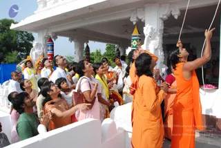 Outdoor photograph of a crowd gathered on a white temple veranda, many looking upward with hands raised or pressed in prayer. In the foreground, two figures in saffron-orange robes stand near the right edge, one raising his arm and pointing toward a white rope or cord that hangs from above, the other with hands folded. Nearby, a man in a green t-shirt and a woman in orange kneel or stand with hands raised. Devotees in white shirts, pink and yellow garments wear yellow shawls. White pillars, a white stone elephant sculpture, and striped multicolored kalasha-like finials decorate the structure, with pink flower petals visible on a tray at right. Trees and a blue tarp appear in the background.