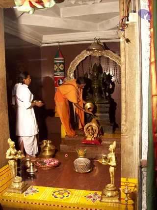 A photograph inside a temple shrine. A man in an orange dhoti and orange upper cloth, with a rudraksha-style mala around his neck, bends forward pouring water from a brass pot over a dark stone deity murti on a granite platform. Behind him a second person in a white dhoti and white shirt holds a small brass vessel. Brass oil lamp figurines stand on either side, a brass kalasha sits with a green coconut-leaf top, a basket holds pink flowers, and a small brass Ganesha figure sits in a red-based niche. A tall green, red and white decorated post and a silver arch frame the shrine. The floor is red stone with a yellow and red painted border.
