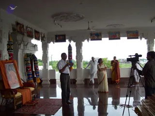Interior hall of a white temple-style building with a glossy floor, carved white pillars and framed pictures along the upper wall. A man in a white shirt and dark trousers stands at center holding a microphone and speaking. Beside him, a woman in a cream sari and another woman in a red-and-gold sari stand near the open window, and a man in white clothes stands at the rail. On the right, a cameraman in a striped shirt operates a video camera on a tripod. At left, a framed portrait of a seated figure with an orange cloth draped over a wicker chair, beside a red mat and tall striped ceremonial poles. No clear basis for identifying a person is visible.