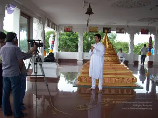 A man in white kurta and white dhoti stands barefoot on a polished marble floor in a white temple hall, holding a handheld microphone and speaking toward a video camera on a tripod operated by a crew member wearing headphones. Beside him stands a tall gilded pedestal in tiered golden form with carved panels. Behind, a seated man in white and a standing man in grey shirt are visible, with a black stone lion, a bell hanging from the ceiling, framed pictures along the pillars and green trees beyond the open arches. Cables trail across the floor.