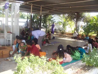 A photograph of an open-sided covered pavilion with a corrugated roof and white pillars, where people sit on the floor on mats and cloths eating from metal plates and bowls, as part of a food distribution. In the centre a man in a white shirt and grey trousers bends forward serving or attending to the seated group. A woman in a purple sari stands at the back near a folding table, and a woman in a light blue top holds a baby on the left. Several children sit among the adults, one girl in an orange dress and another with flowers in her hair. Green plants and palm trees surround the pavilion, with stacked white plastic chairs on the left and red-brown bricks at the lower left.