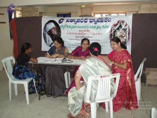 A photograph of a free medical camp indoors at a hall. Four women sit at a white folding table covered with paper, with a woman in a blue top and dark patterned sari writing on a form. Behind them hangs a white banner with Telugu text and a circular photo of a bearded figure in white headwear, plus an image of a decorated deity murti with garlands. On the left a woman in a black and blue floral outfit sits on a white plastic chair, and a woman in a magenta sari sits in the center. A woman in a red sari with gold border sits at right, and a woman in a grey-green sari with a pink hair clip sits in front. Cardboard boxes are stacked along the wall at right.