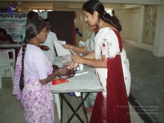 A photograph of a free medical camp in a hall with tiled floor. A woman in a white saree with red border and gold bangles stands at a white table, handing medicine or a paper to an older woman in a lavender floral dress and white blouse with a long grey braid, who holds a red packet and a brown handbag. Behind the table a second worker in a grey saree and another person in white stand near a brown screen, and a banner with Telugu text hangs on the wall. Small bottles, a plastic bag and printed forms sit on the table.