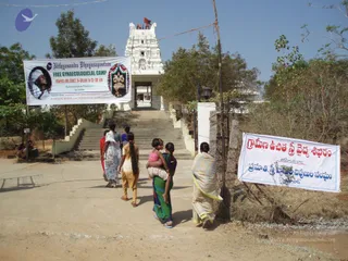 A photograph of a temple entrance with a white gopuram-style tower topped by a red flag. A banner on the left, headed Nithyananda Dhyanapeetam, announces a Free Gynaecological Camp, March 4th 2007, 8:30am to 12:30pm, with a portrait of a bearded man in white head cloth and a deity image. A Telugu banner on the right reads a community health camp. Several women and girls, one carrying a child on her hip, walk up a concrete staircase toward the temple gate under a clear sky. One seated figure sits at the left edge.