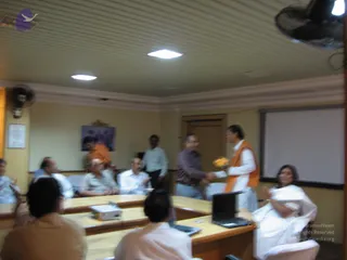 Indoor meeting room with cream walls, a ceiling fan and tube lights, and rows of wooden tables with laptops and a projector. A man in white kurta pyjama with an orange shawl stands at right holding a bundle of yellow flowers and shaking hands with a man in a brown shirt. Several seated men in white and light shirts watch, and a woman in white sari sits at right applauding. A small framed picture hangs on the back wall.