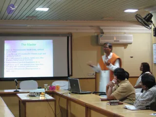 A classroom-style presentation room with wood-panelled desks, a projector screen, and ceiling fans. A standing man in a white dhoti and an orange sleeveless vest gestures while speaking toward seated listeners. The projected slide, titled "The Master", lists bullet points about a teacher's early life and founding of an institution. Seated listeners include a man in a tan shirt with his chin resting on his hand, a man in a checked shirt at a laptop, and a woman in white at the right. A glass of water, flowers and a laptop sit on the desk.