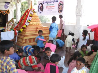 A group of children sit and kneel on a polished marble floor in a temple pavilion, many holding pink and red paper sheets and drawing or crafting with pencils. Behind them a large golden stepped pyramid-shaped stand, decorated with red, white and yellow flower garlands, sits beside a framed picture on a draped table. A white-pillared hall with an orange marigold garland overhead forms the background. A hanging banner shows a portrait of a bearded man in a white turban beside a deity image, with a printed title. Several children wear blue school-style shirts, others wear striped shirts, pinafores and frocks.