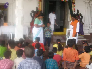 A photo of children seated on the floor in front of a temple shrine, facing a small stage. A woman in a white sari with a light teal blouse stands at left with hands together, and a young boy in a blue-green checked shirt stands beside her with palms joined. At right, a man in white clothes and an orange shawl stands by a white chair, holding a small object. Behind them, a man in white stands in the temple doorway, and two dark deity murti with garlands are visible at the sides. Several children in bright yellow, orange, pink and checked shirts sit in the foreground.